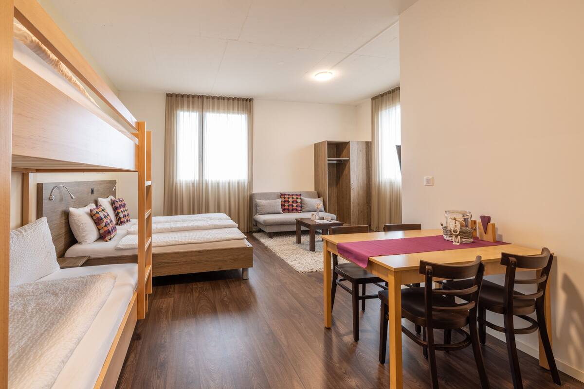 Quarto de hotel moderno com cama de madeira, sofá, mesa de jantar com cadeiras e uma pequena cozinha.