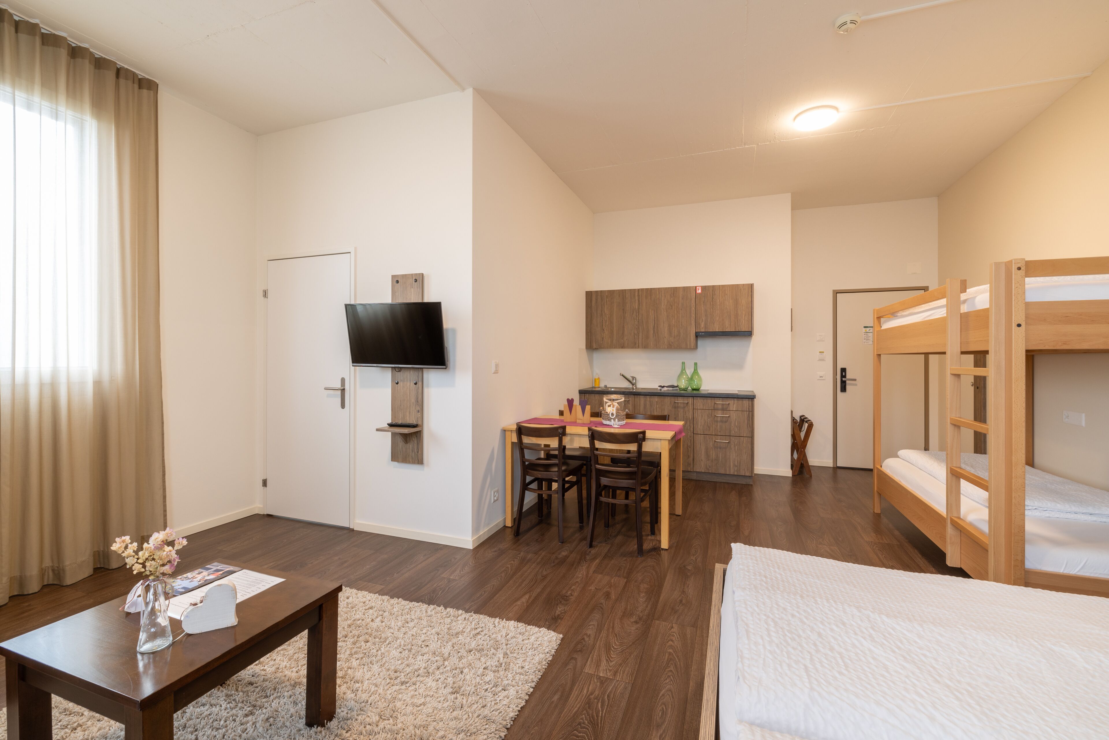 A well-lit room with a wooden dining table, chairs, a small kitchen area, a bunk bed, and a TV mounted on the wall.