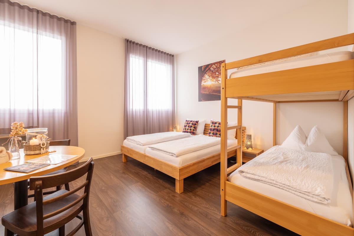 Quarto com beliche, escrivaninha e cadeira. Há uma janela com cortinas, um quadro na parede e uma mesinha com um candeeiro e um vaso.