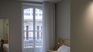 Habitación de hotel con una cama grande, un escritorio de madera, una mesita de noche y un ventanal con vistas a edificios.