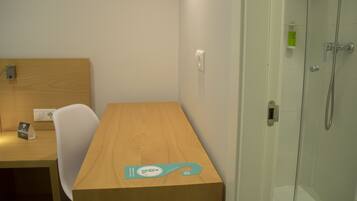 Habitación de hotel con escritorio de madera, una silla, una mesita pequeña, cabina de ducha de vidrio y un inodoro blanco.