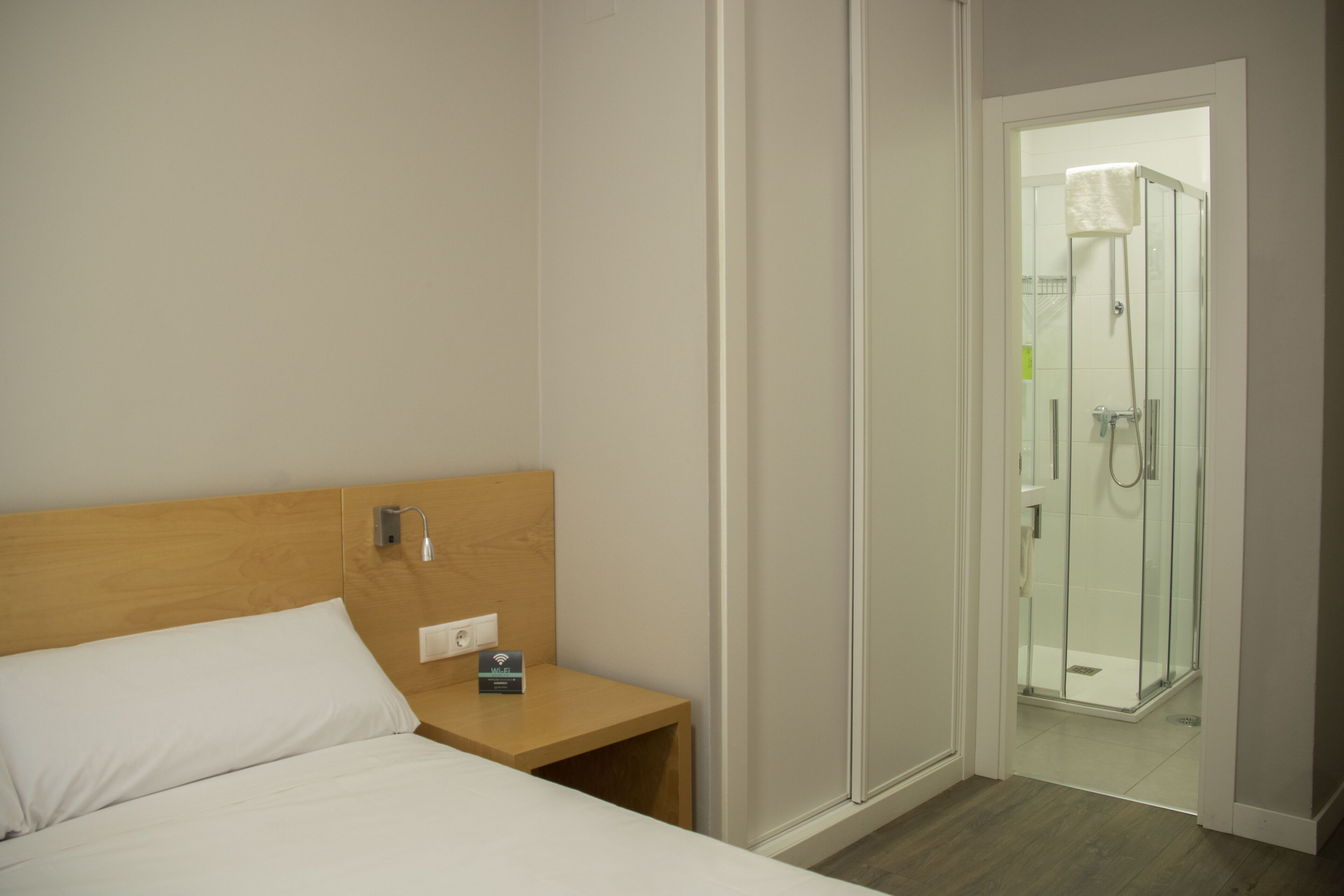 A hotel room with a wooden headboard, a single bed, a nightstand with a power outlet, and a glass-enclosed shower area.