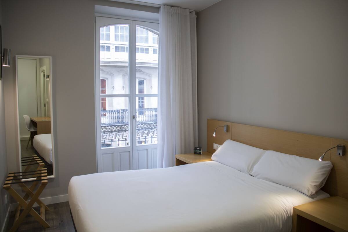 Habitación de hotel con cama grande, mesita de noche, espejo y ventana con vista a un edificio y balcón.