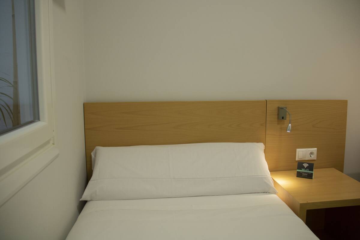 Una cama bien hecha con sábanas blancas en una habitación con cabecera de madera. Se observa un pequeño mesito de noche con el símbolo de Wi-Fi y un enchufe.