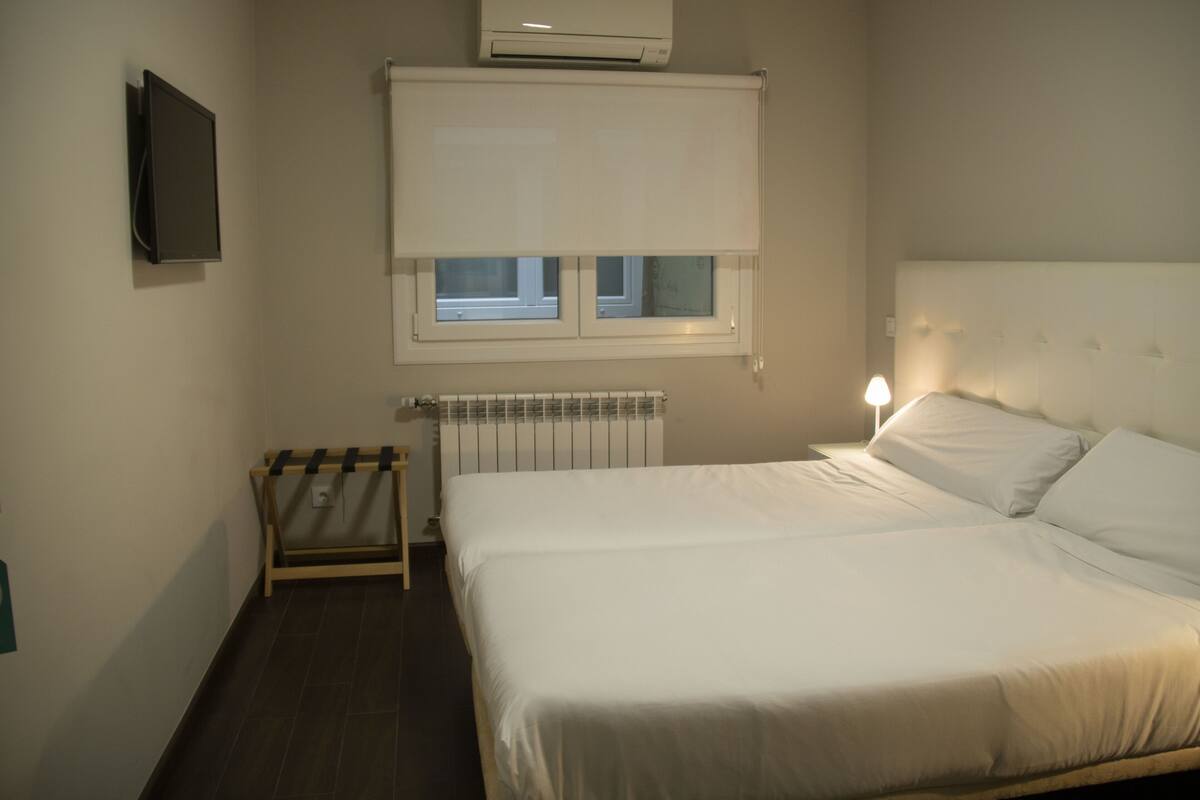 Habitación de hotel con cama, ventana con persianas, radiador, mesita de noche y televisor montado en la pared.