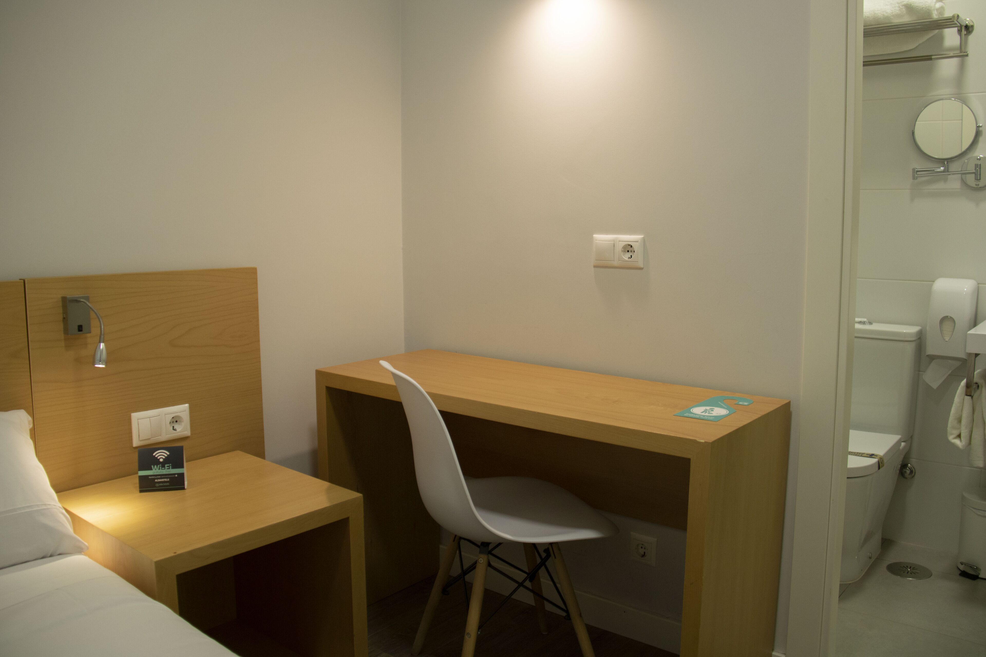 Ein Hotelzimmer mit einem Holztisch, einem weißen Stuhl, einem Bett, einem kleinen Tisch mit einem WLAN-Symbol, einer Steckdose und einem Badezimmer mit Toilette und Spiegel.
