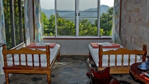 Une chambre avec deux lits en bois, un sol en pierre et une vue sur la verdure et les montagnes, grâce à de grandes fenêtres.