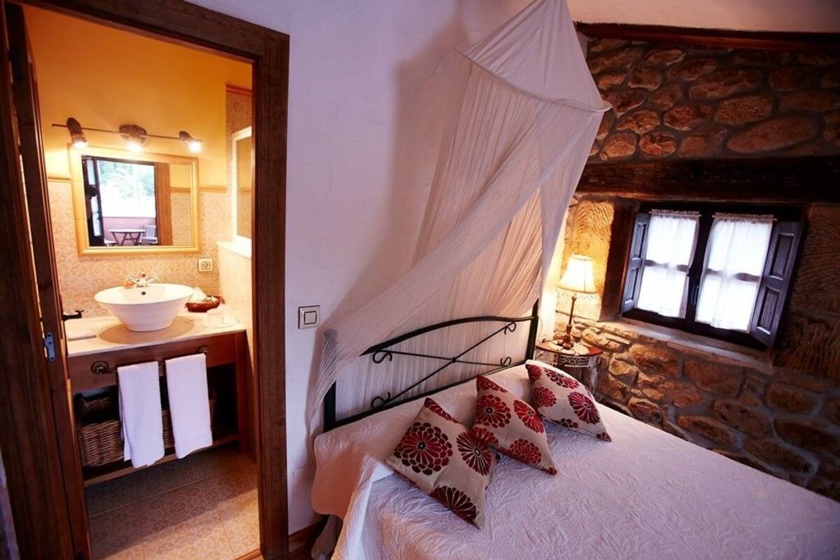 A bedroom with a stone wall, a bed with a canopy, a bedside table with a lamp, and a bathroom visible through an open door.