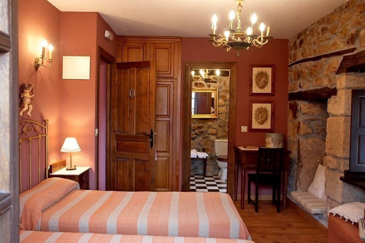 A bedroom with a bed, a chandelier, a wooden wardrobe, a stone wall, and a mirror.