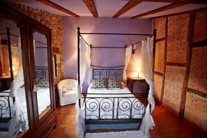 Un lit à baldaquin avec une structure en métal et une literie blanche, un fauteuil blanc et un mur orné d’un motif en briques.