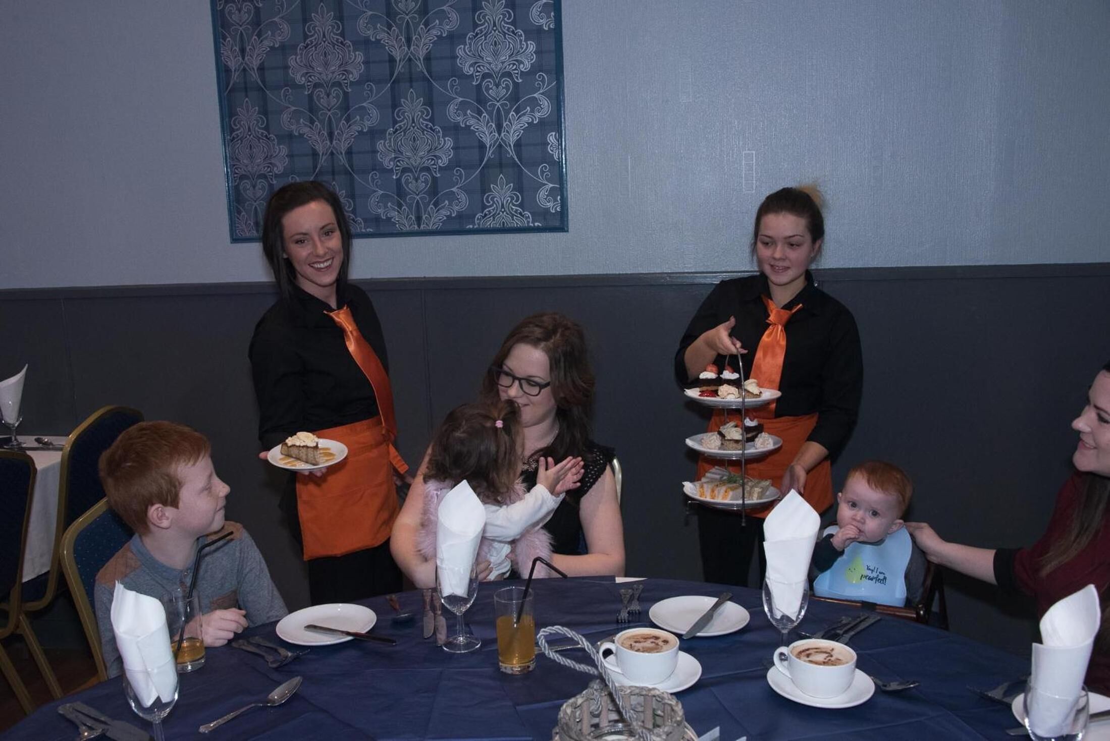 A group of people seated around a table with a blue tablecloth, plates, cups, and a glass of liquid.