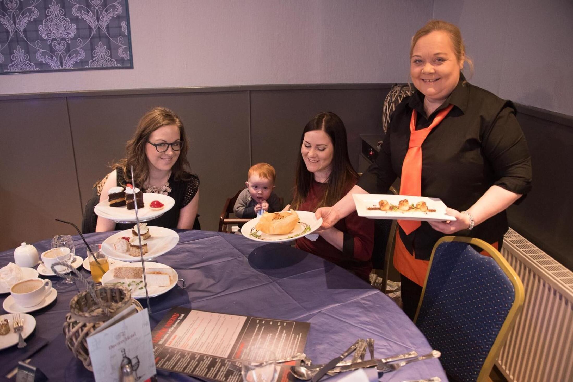 Un groupe de personnes est assis autour d’une table recouverte d’une nappe bleue, avec des assiettes remplies de nourriture et un panneau indiquant le menu.