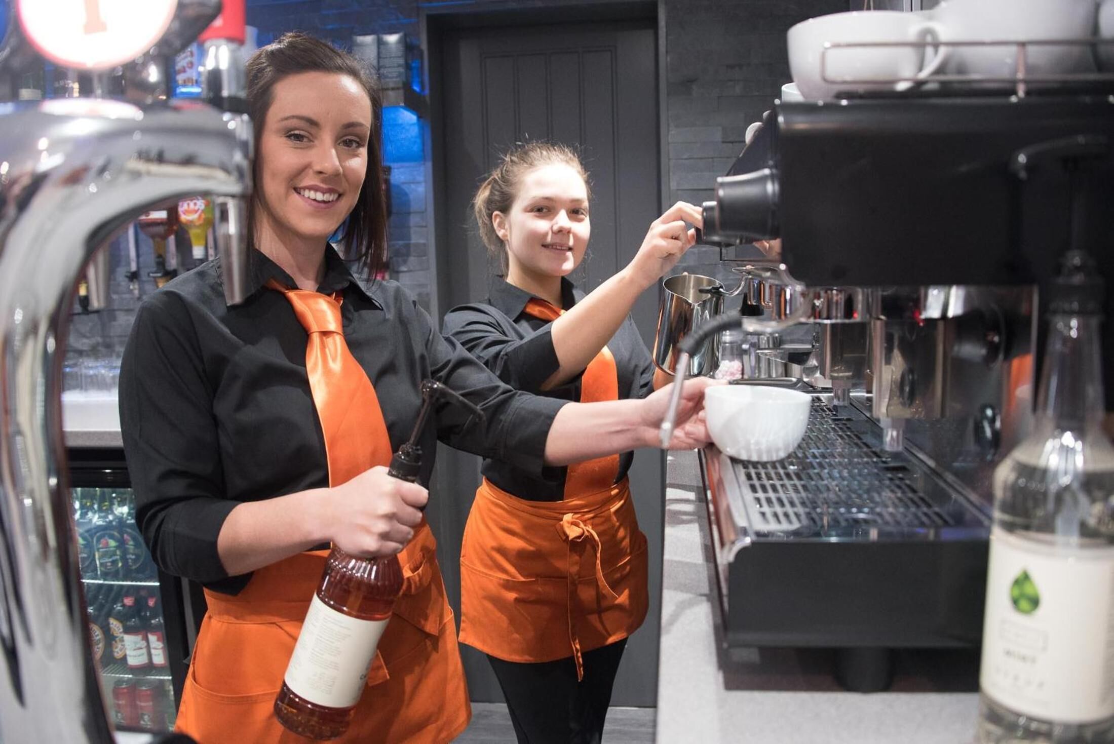Two baristas in an indoor setting, one holding a bottle of syrup and the other pouring into a cup.
