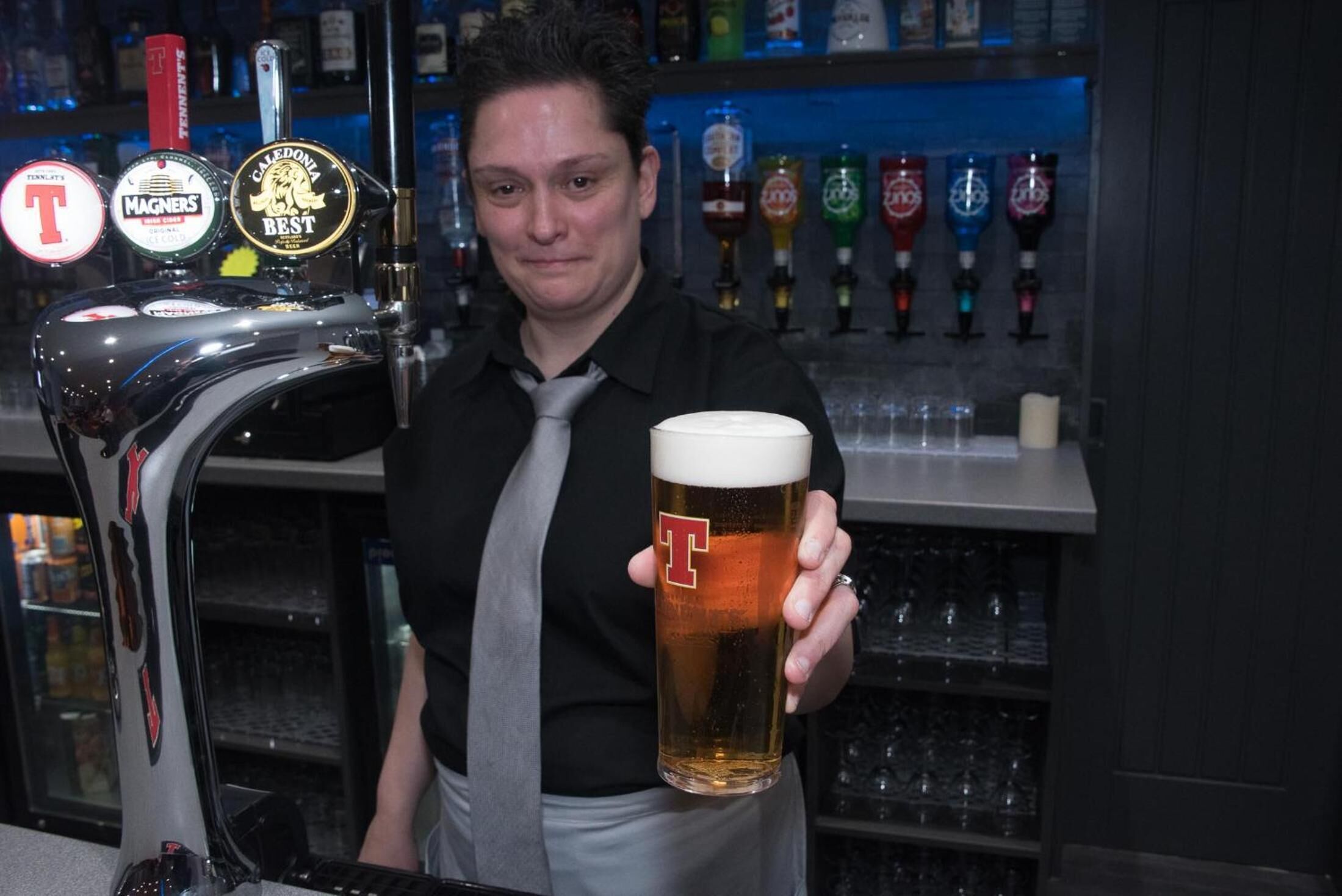 A bartender holding a beer mug with a logo, behind a tap with multiple beer brands.