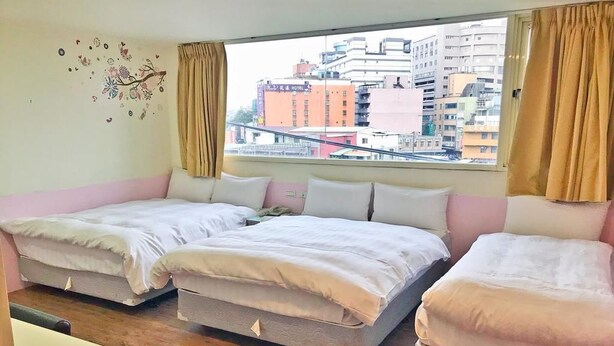 A hotel room with two beds, a small wooden table, and a view of buildings through the window. - Happy Holiday Hotel (Taoyuan City)