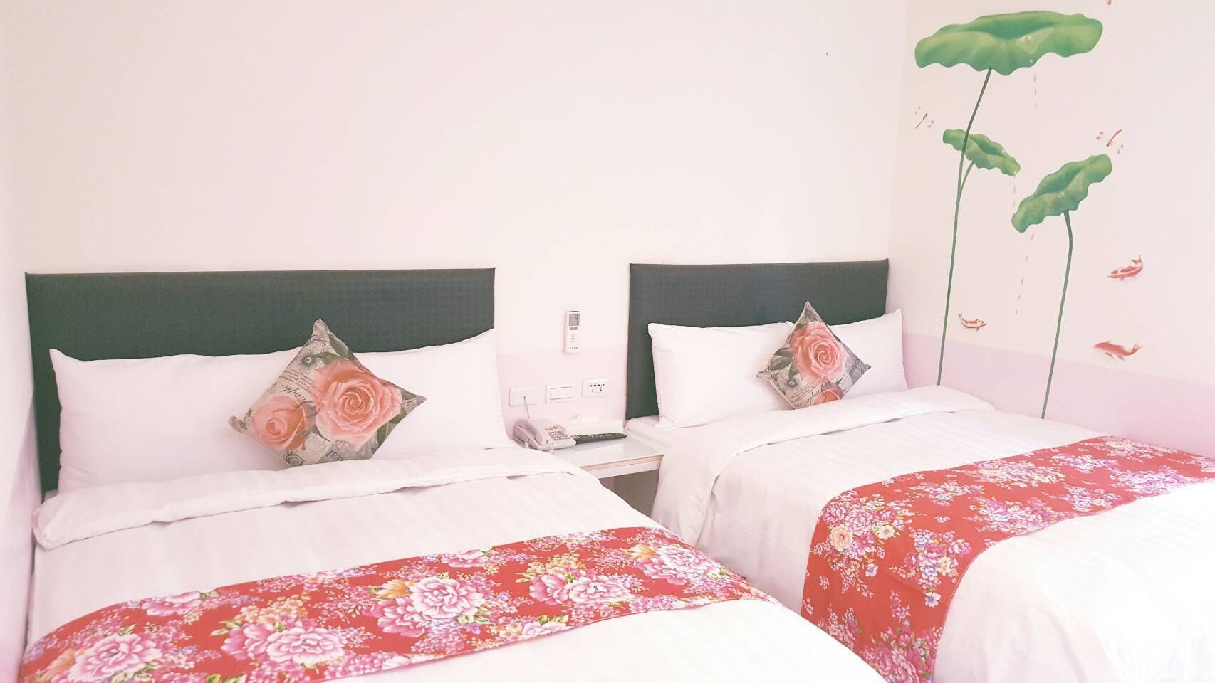 A hotel room with two beds, each with a floral bedspread and a green headboard. There is a nightstand with a phone and a lamp between the beds.