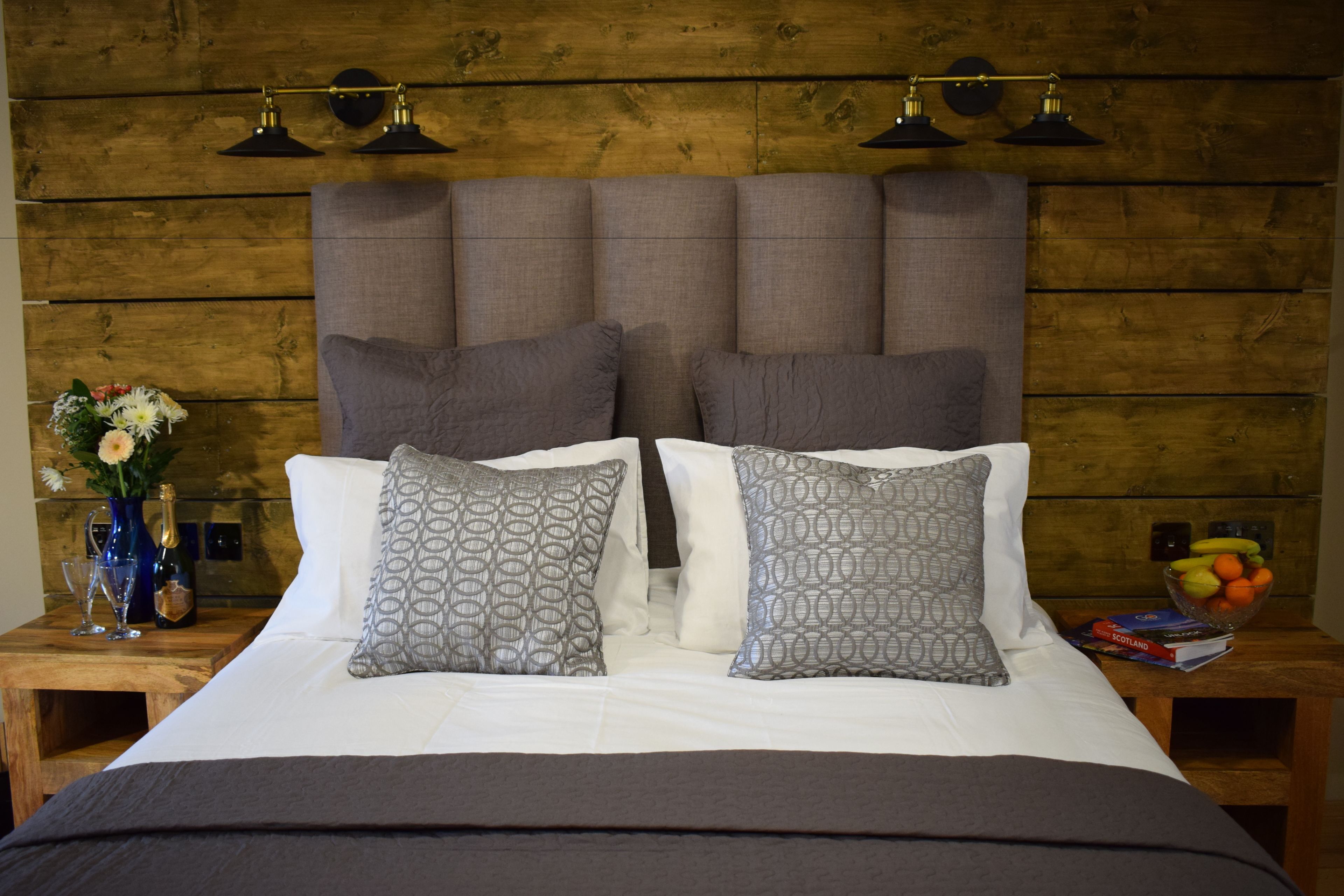 A bed with a wooden headboard, two patterned pillows, a white comforter, and a dark blanket. There are two bedside tables with a vase of flowers and a bowl of fruit.