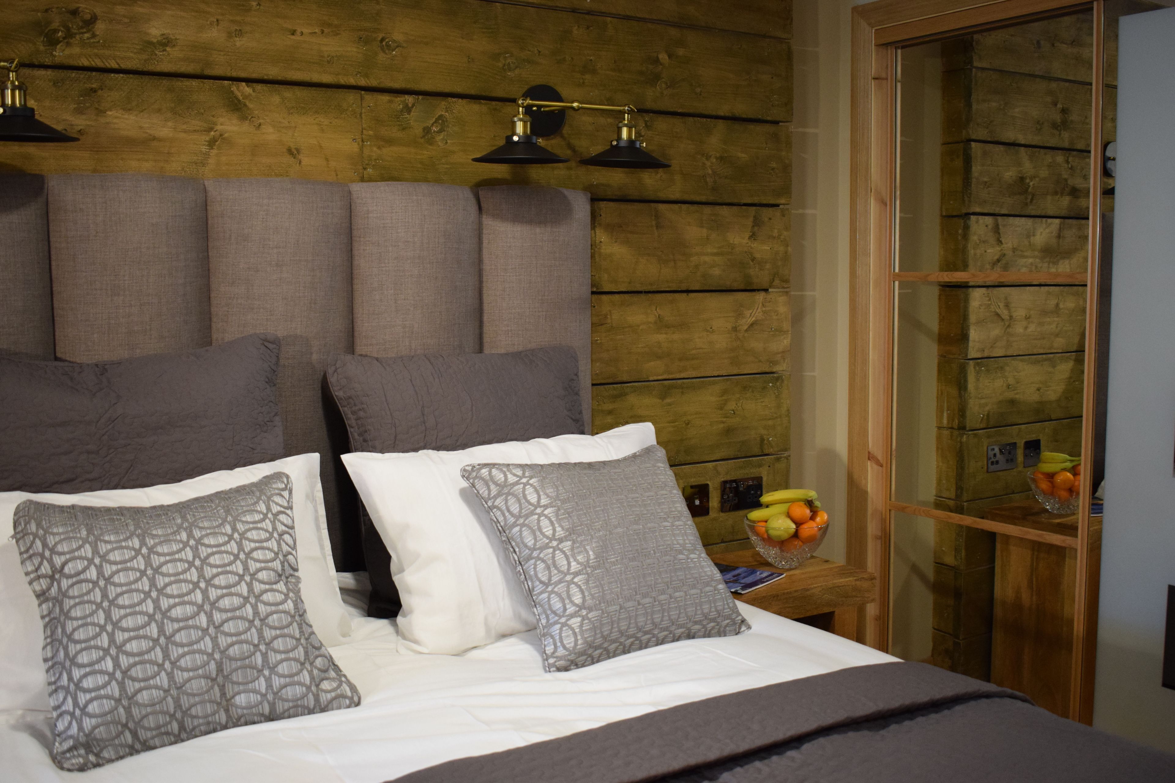 A bed with grey and white pillows, a wooden headboard, and a nightstand with a fruit bowl.