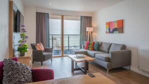 A modern living room with a grey sofa, a wooden coffee table, and a red armchair. There is a large window with curtains, a flat-screen TV mounted on the wall, and a colorful abstract painting above the sofa. - Talbot Suites at Stonebridge (Wexford)