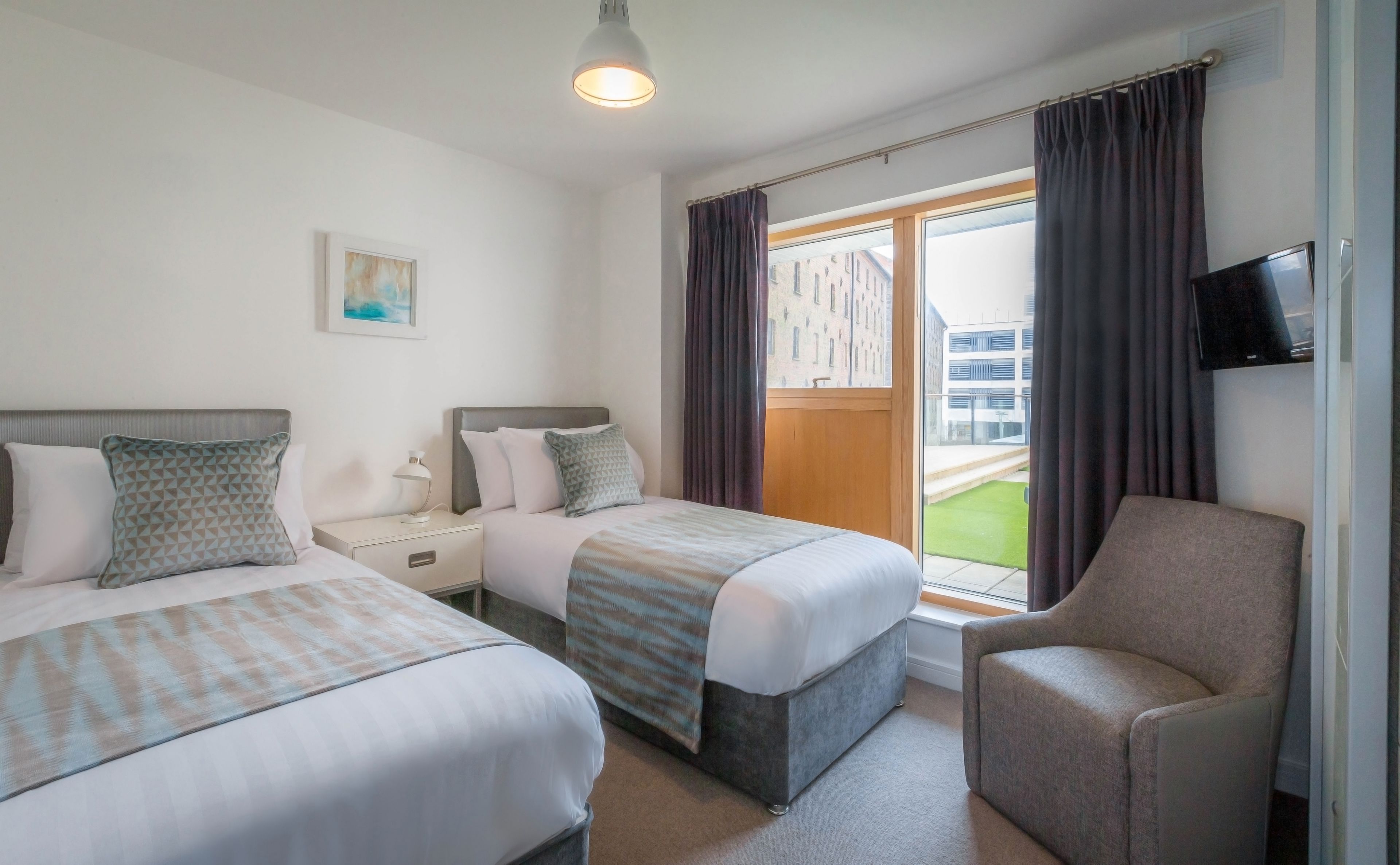 A hotel room with two beds, a chair, a TV, and a window with curtains.