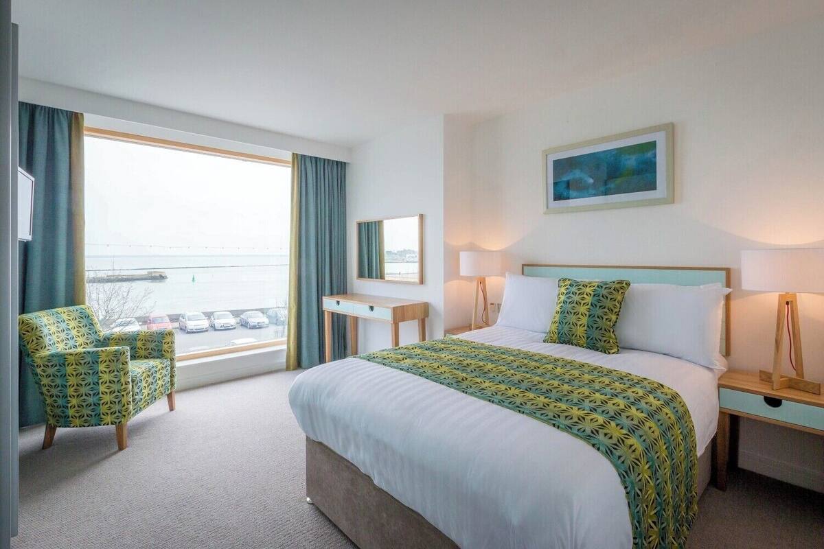 Habitación de hotel con cama, escritorio, silla, ventana con vistas y un cuadro en la pared.