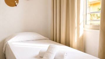 Una cama individual con sábana blanca y dos toallas, una ventana con cortinas y dos adornos en la pared.