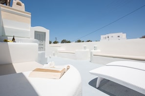 Une pièce aux murs blancs, avec un coin lecture baigné de lumière, une petite table sur laquelle repose un livre et un banc.