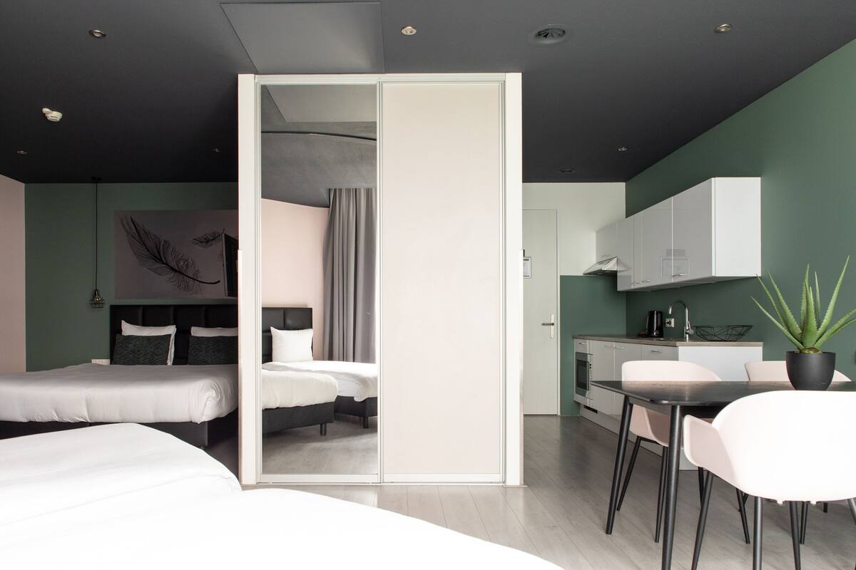 A modern hotel room with a bed, bedside tables, a desk, and a chair. There is a kitchen area with cabinets and a sink.