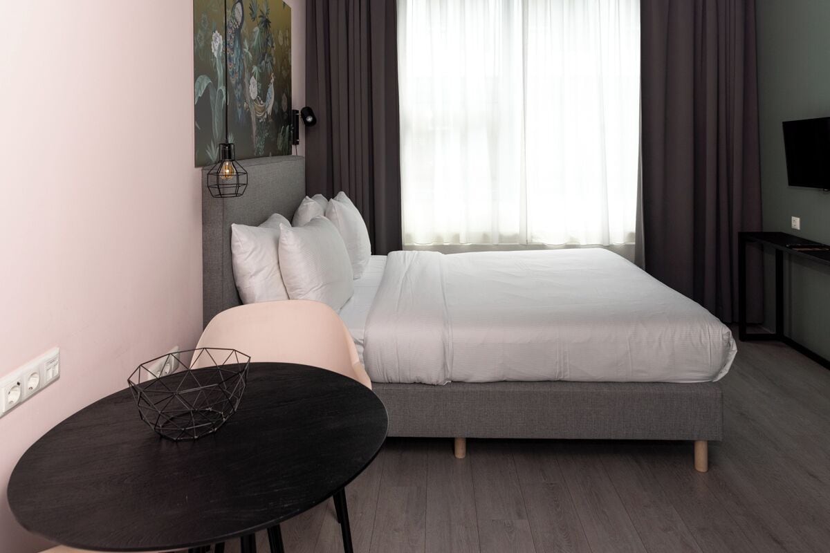 Un dormitorio moderno con una cama, una mesita redonda con una cesta de mimbre, un cuadro en la pared y un televisor sobre un mueble bajo.