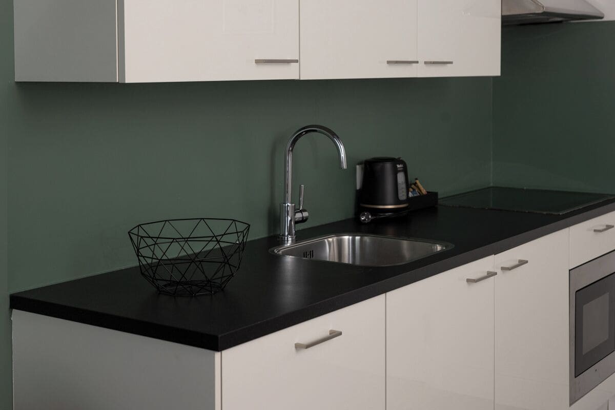 Una cocina moderna con encimera negra, armarios blancos, un lavamanos de acero inoxidable y una cafetera.