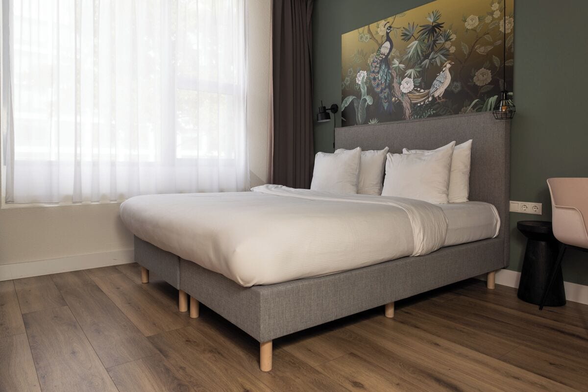 Un dormitorio moderno con una cama gris, ropa de cama blanca, un ventanal con cortinas translúcidas y un cuadro en la pared de pájaros y flores.