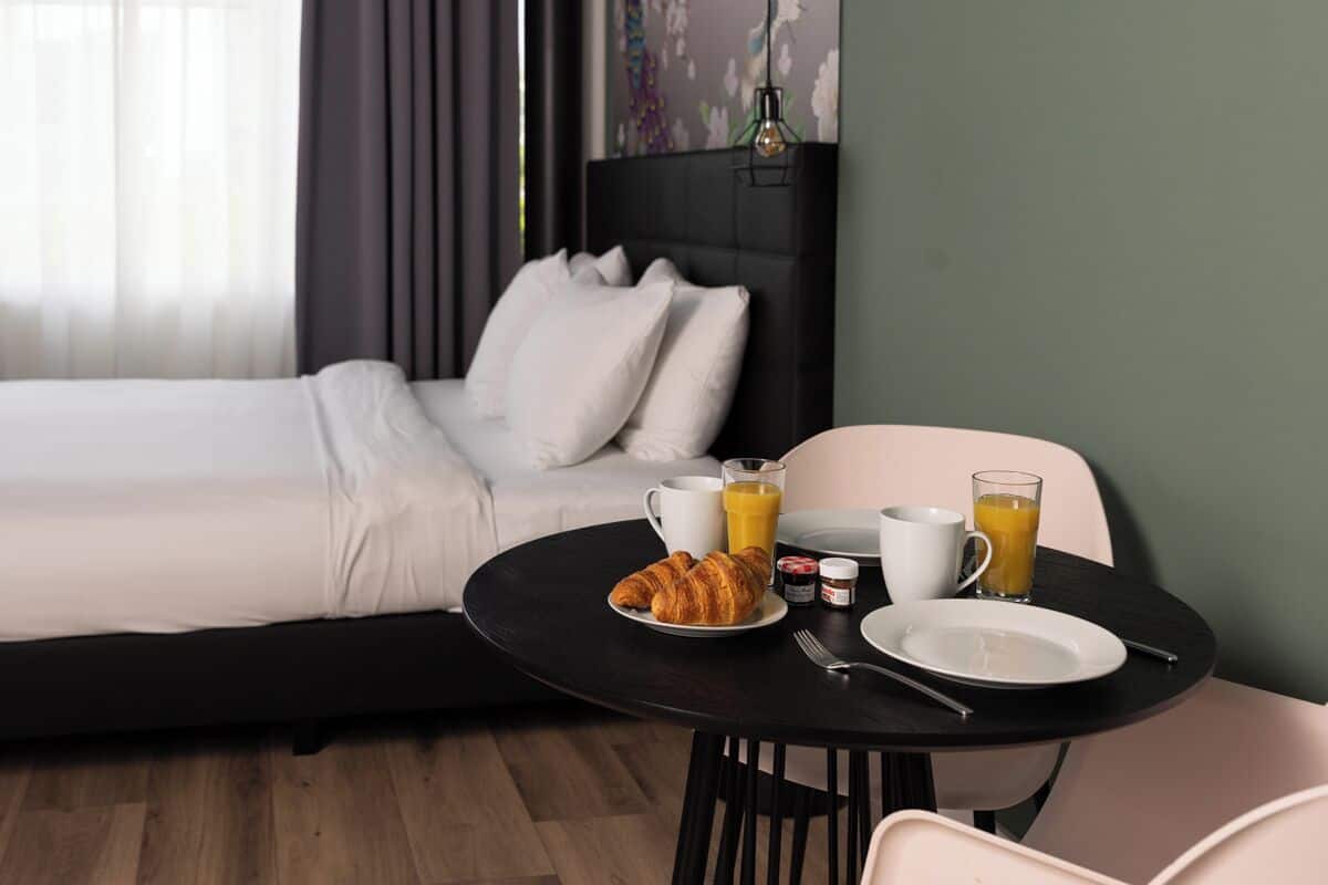 A hotel room with a bed, a breakfast table with croissants, juice, and a coffee cup.