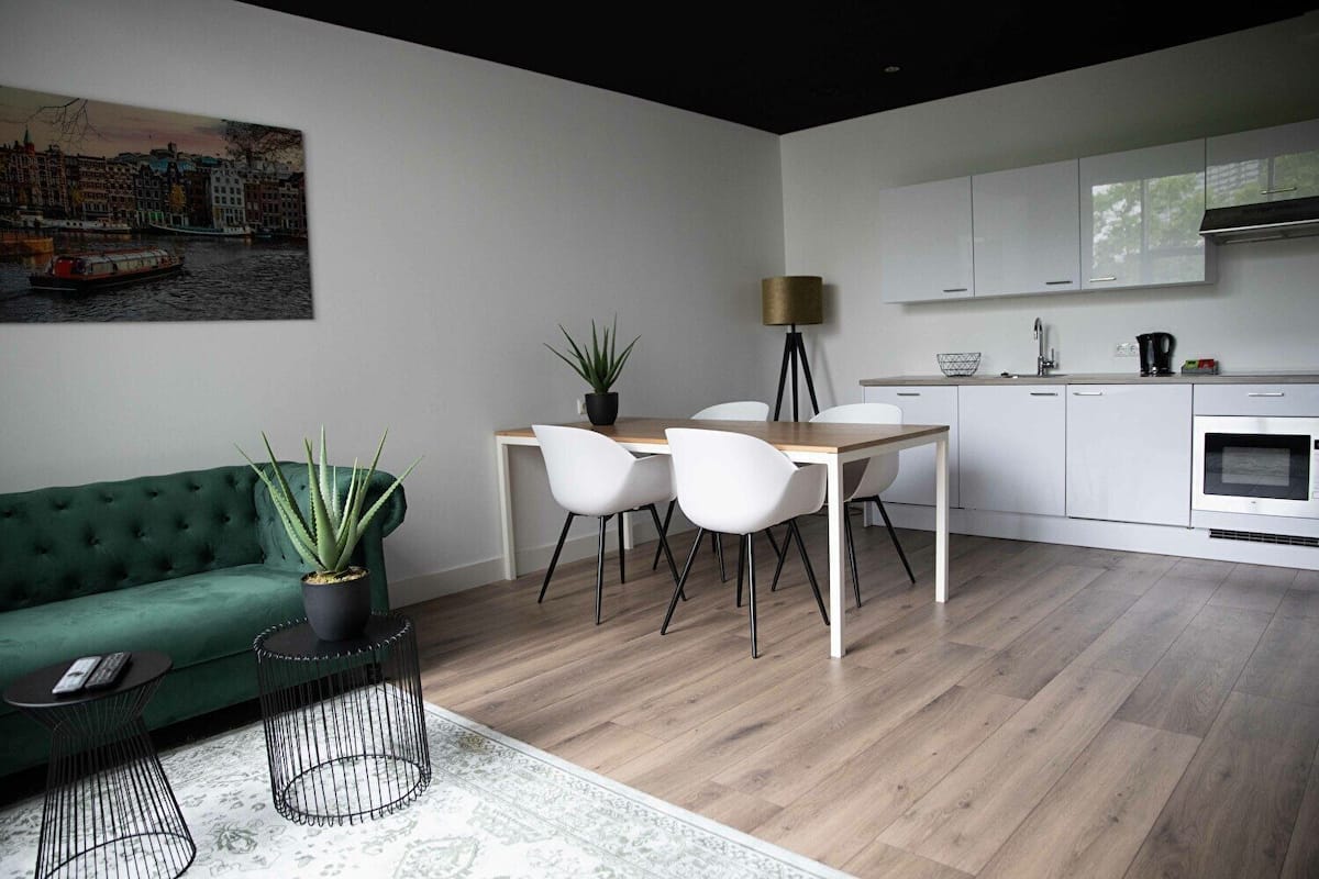 A modern living room with a green sofa, a dining table with white chairs, a kitchen with white cabinets, and a painting of a cityscape.