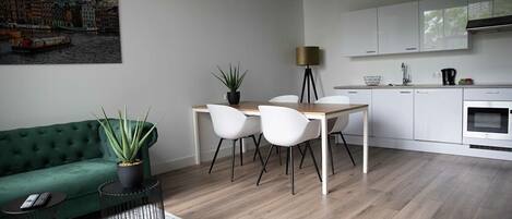 A modern living room with a green sofa, a dining table with white chairs, a kitchen with white cabinets, and a painting of a cityscape.
