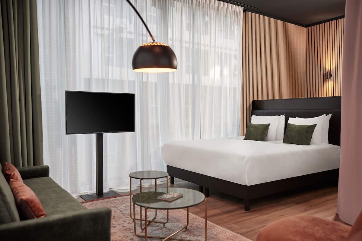 A modern hotel room with a large bed, a sofa, a small table, a TV, and a large window with sheer curtains.
