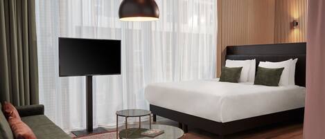 Habitación de hotel moderna con una cama grande, un sofá, una mesita, un televisor y un ventanal con cortinas translúcidas.