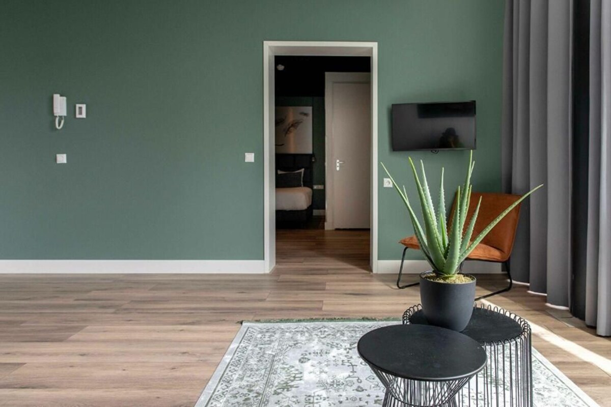 A modern living room with a green wall, a wooden floor, a round black table, a potted plant, a TV, and a door leading to another room.
