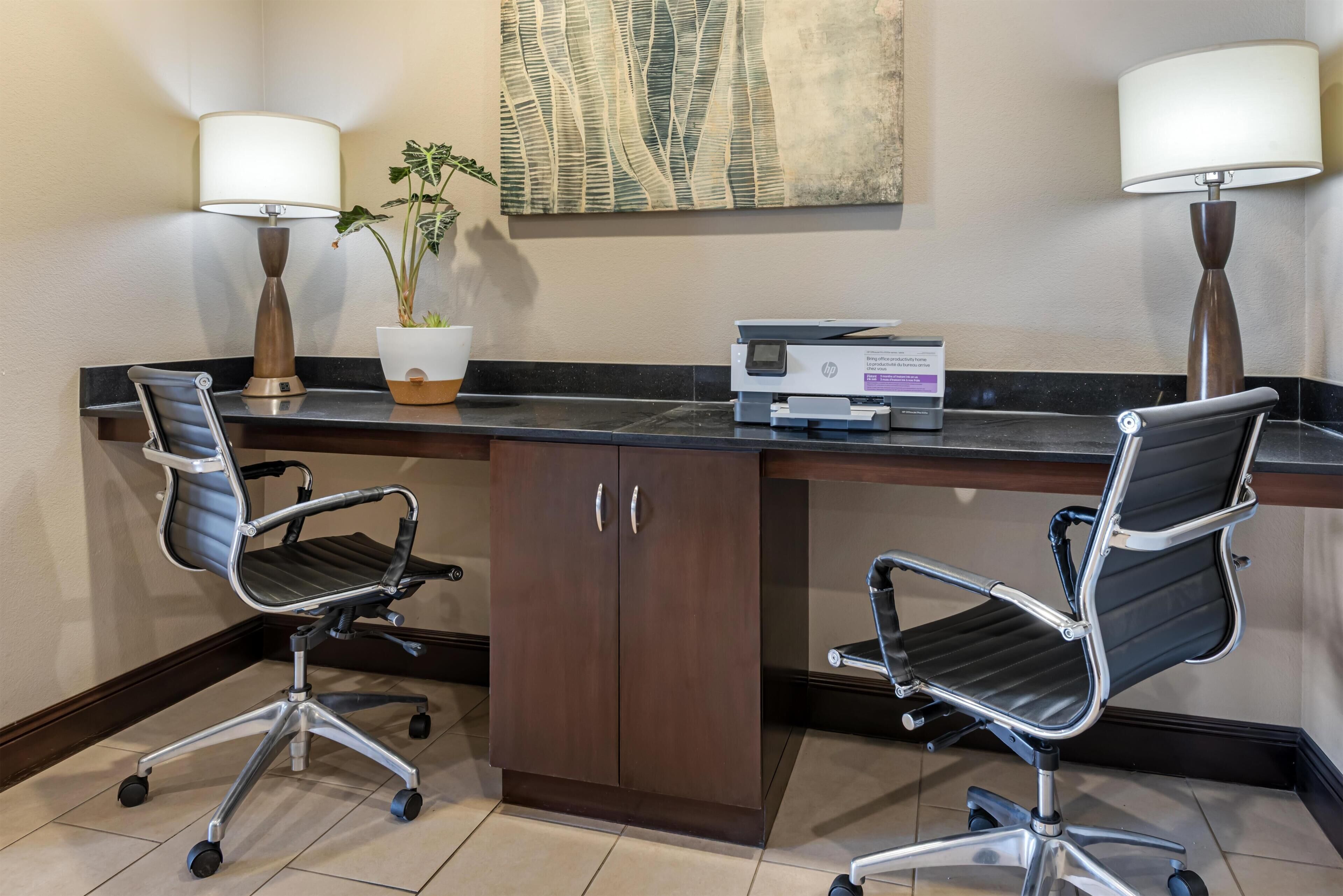 A hotel room with a desk, two office chairs, a printer, a potted plant, and two lamps.