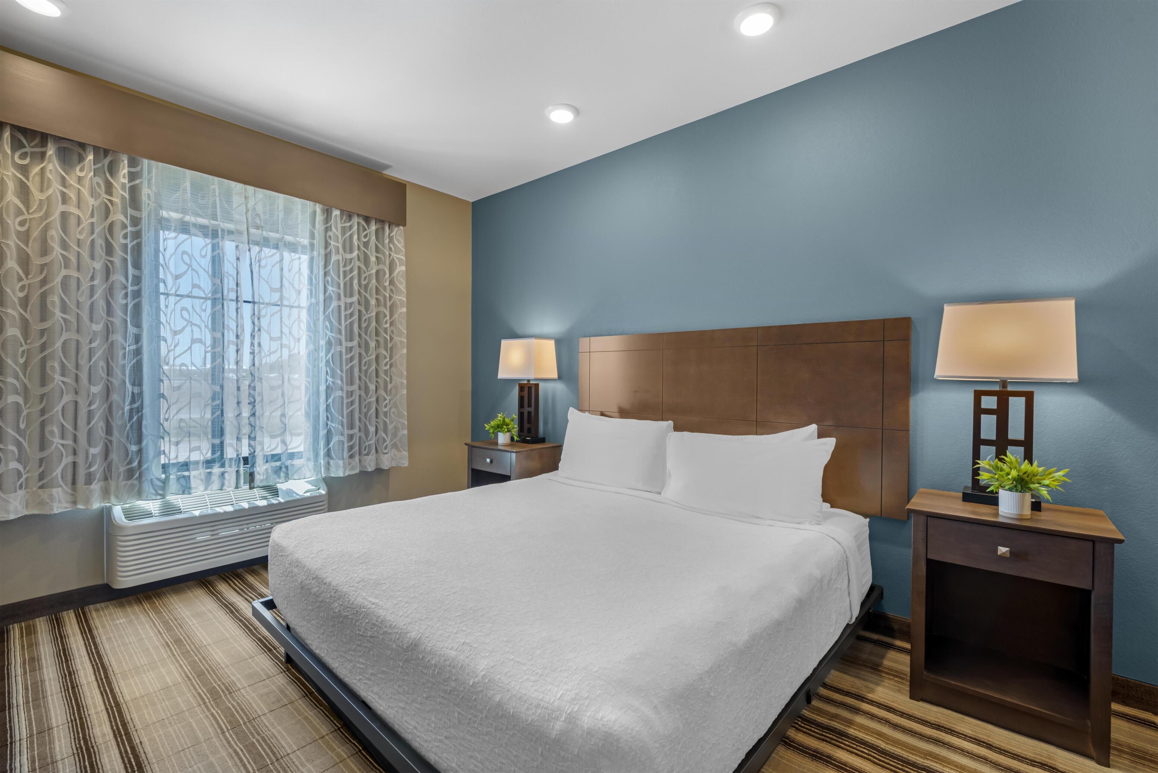 A hotel room with a large bed, two bedside tables with lamps, a window with curtains, and a wall-mounted heater.
