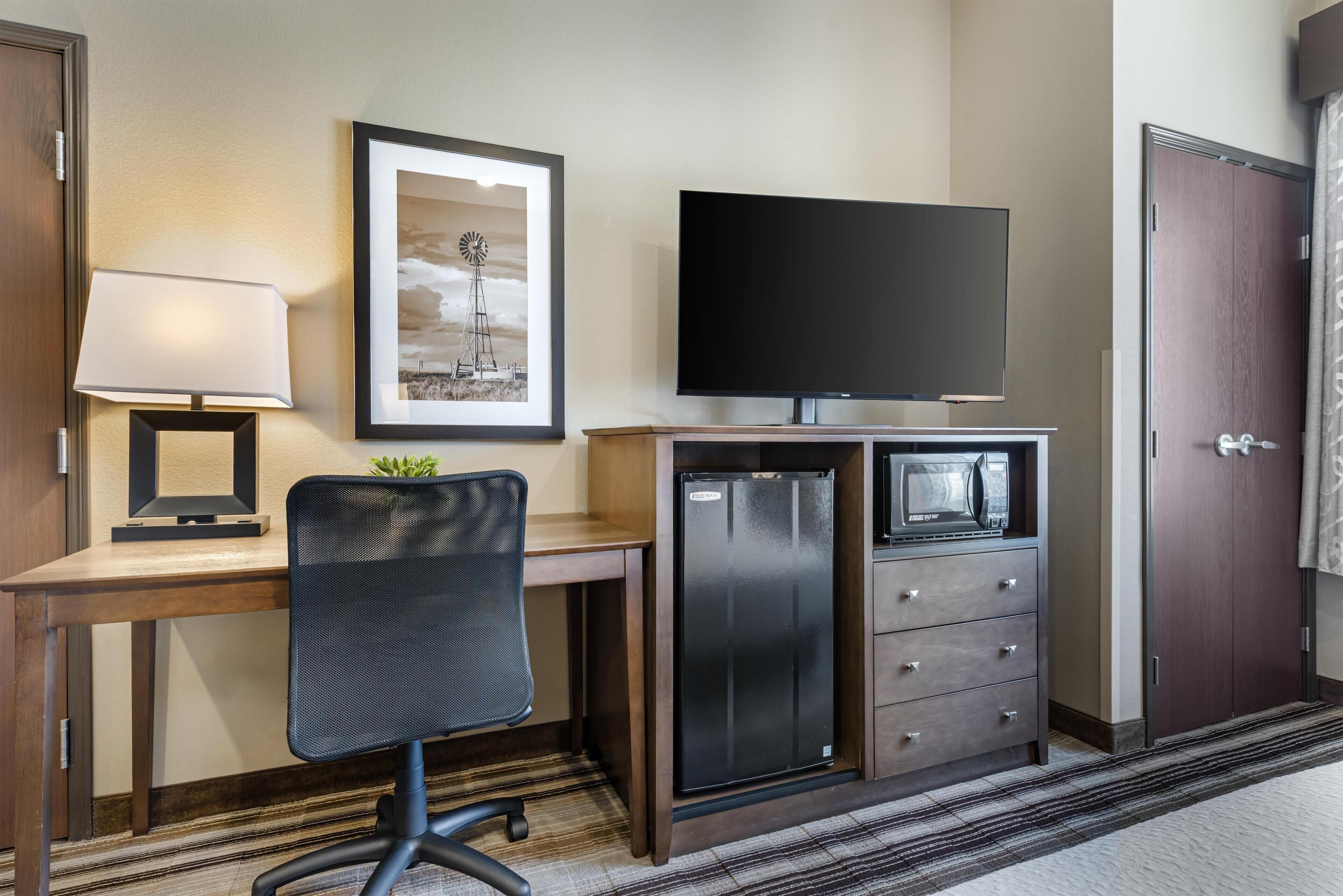 A hotel room with a desk, chair, television, refrigerator, microwave, and a door.