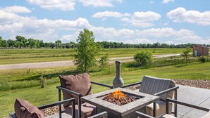 Une terrasse dotée de mobilier d’extérieur et d’un foyer, offrant une vue sur un champ verdoyant et une route.
