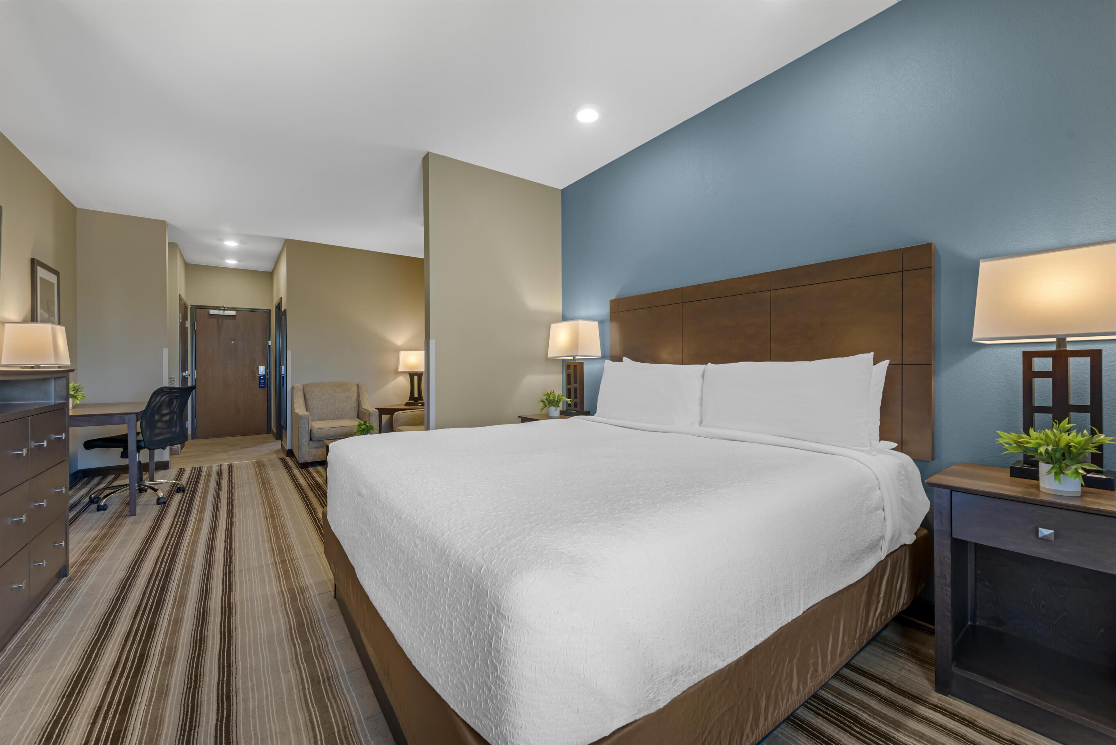 A hotel room with a large bed, a desk, a chair, and a nightstand with a lamp.