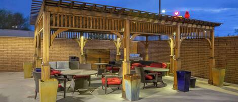 An outdoor patio with wooden pergola seating, tables, and chairs, and a brick wall in the background.