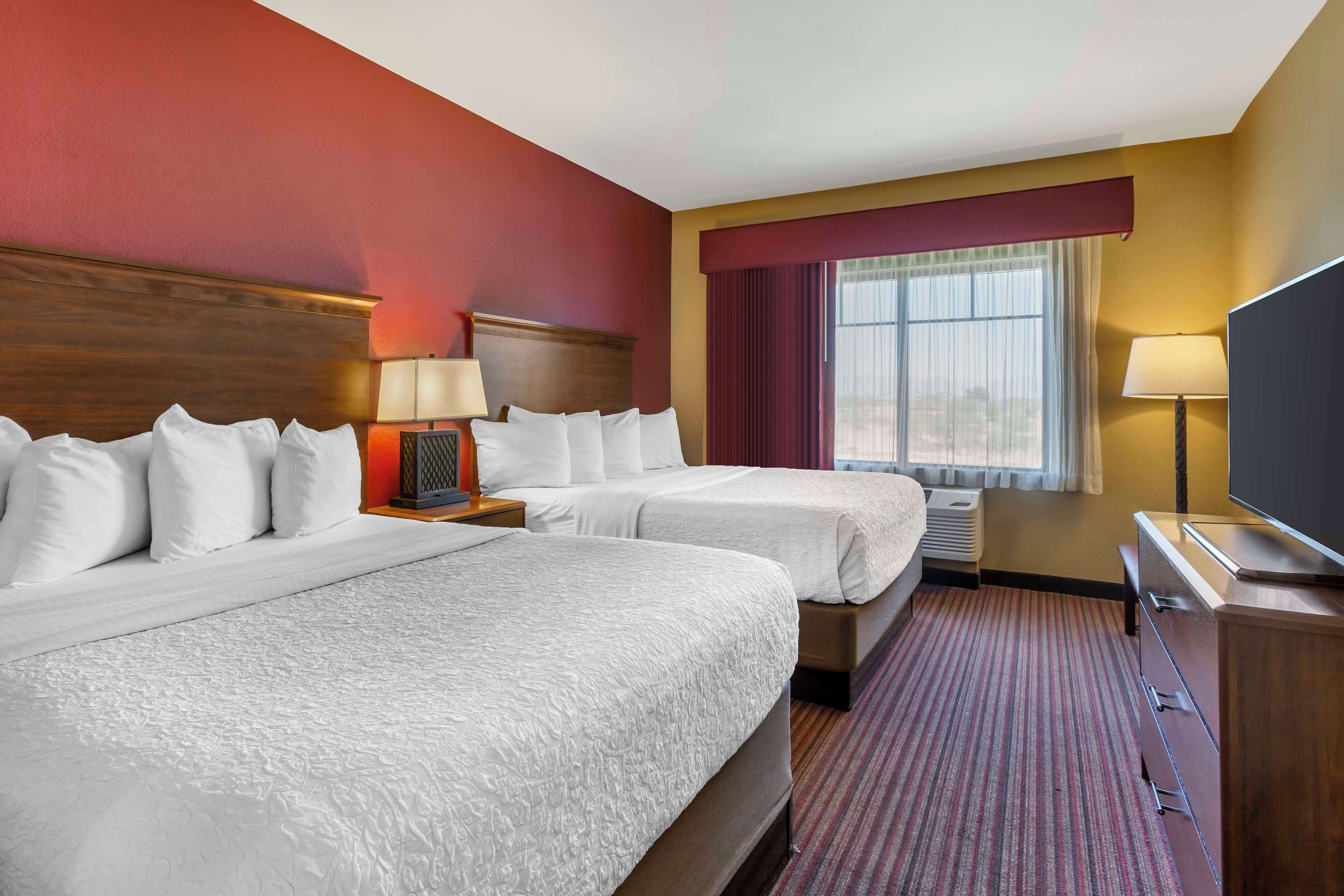 Quarto de hotel com duas camas, televisão, escrivaninha com candeeiro e janela com cortinas.
