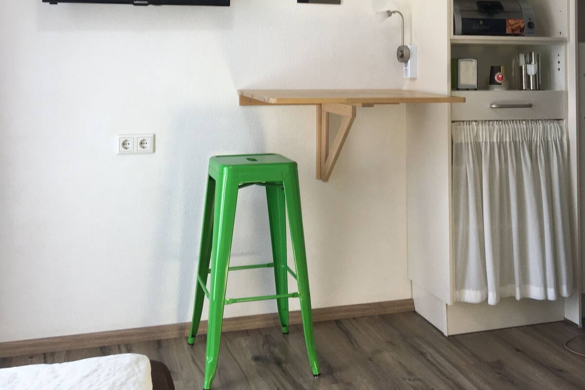 Una habitación pequeña y funcional con televisión, reloj y un escritorio de madera con taburete.