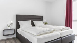Un dormitorio moderno con un cabecero oscuro, ropa de cama blanca, una mesita de noche pequeña con una lámpara y una planta en maceta.
