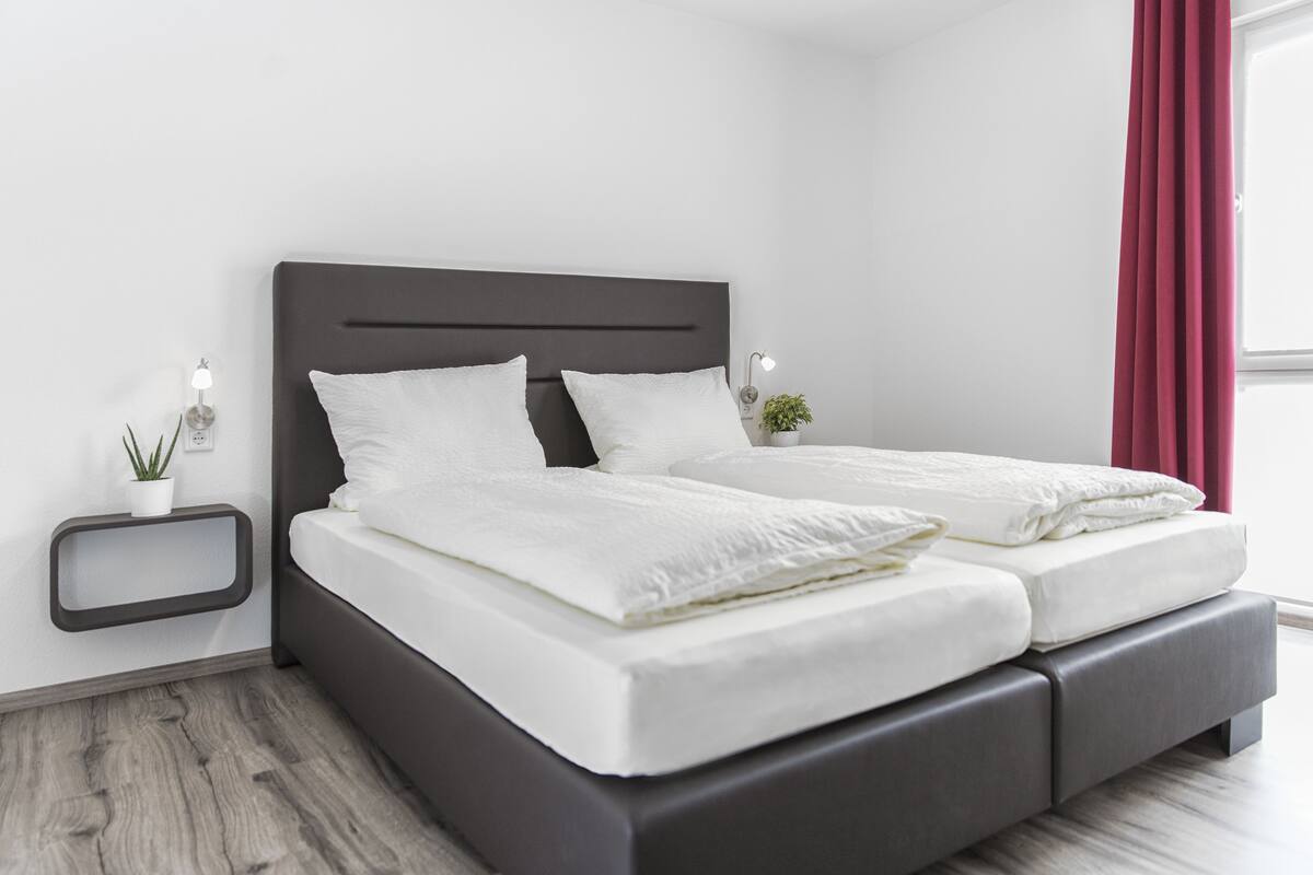 Un dormitorio moderno con un cabecero oscuro, ropa de cama blanca, una mesita de noche pequeña con una lámpara y una planta en maceta.