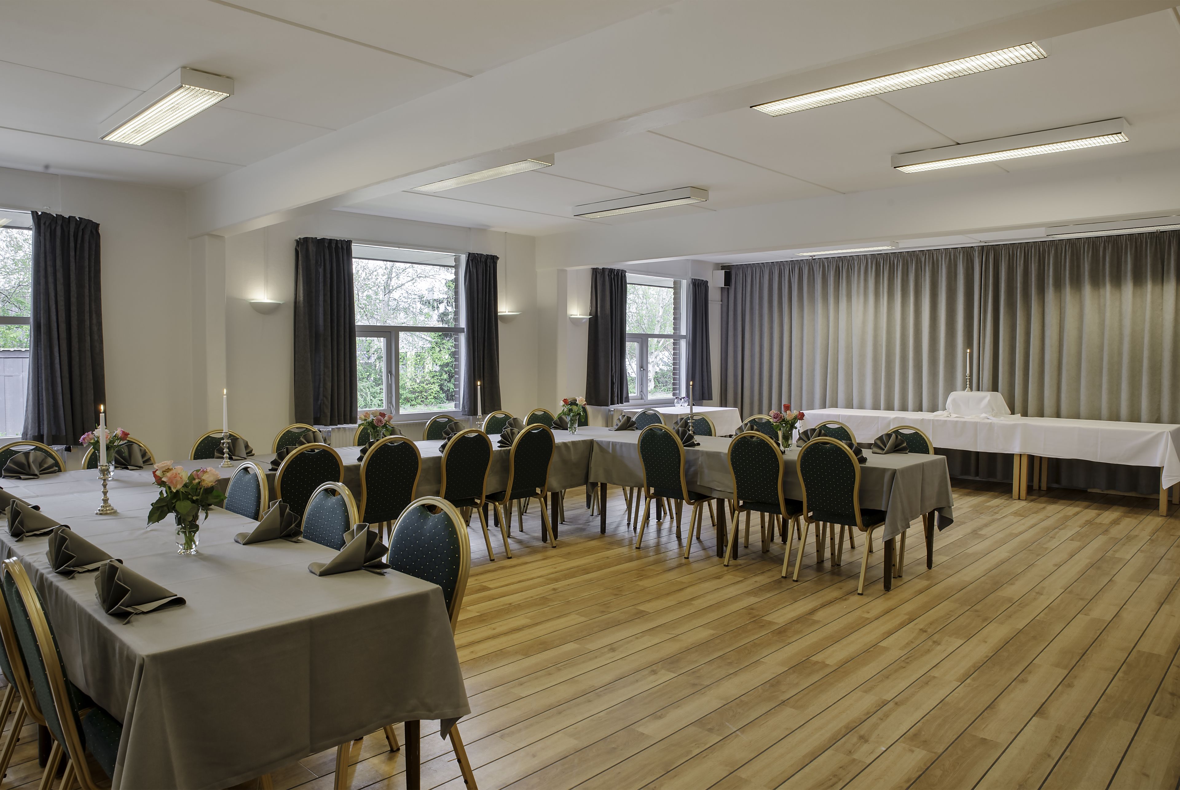 A conference room with multiple tables and chairs, large windows, and a long table covered with a white tablecloth.