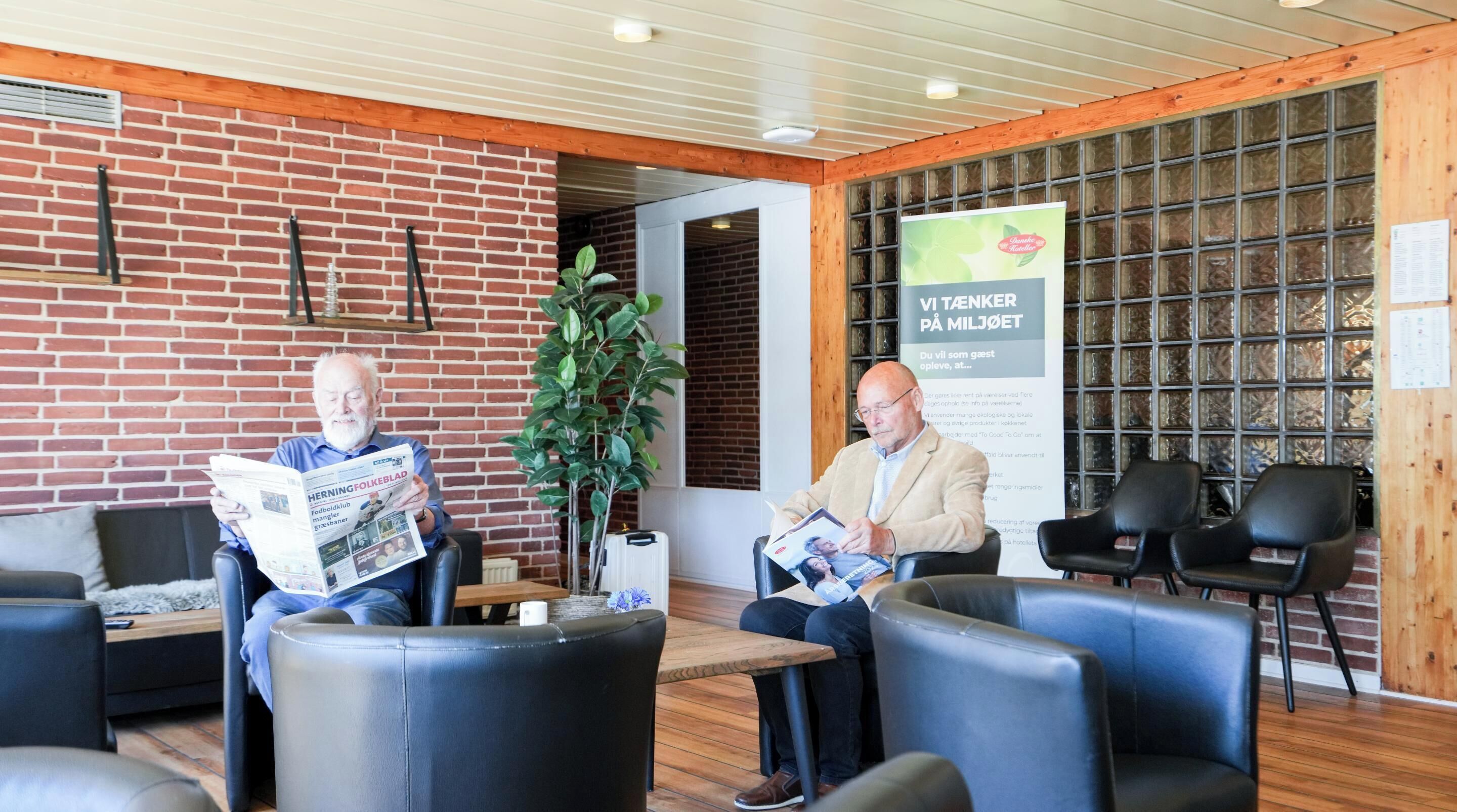 Two men seated in a modern lobby with brick walls, wooden accents, and a large informational poster.
