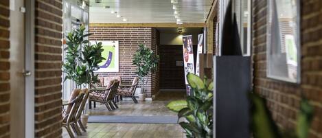 A hallway with a brick wall, wooden floor, and potted plants. There is a seating area with chairs and a table in the distance.